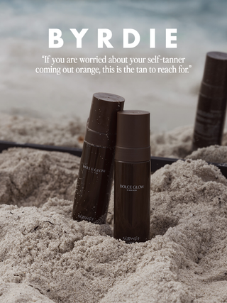 Three dark brown Dolce Glow self-tanner bottles standing in sand, with beach water blurred in the background and the text 'BYRDIE' 