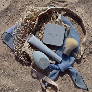 Dolce Glow Travel Bundle products including SPF50 Body Balm, tanning contour, brush, and blending sponge on a sandy beach with mesh bag.